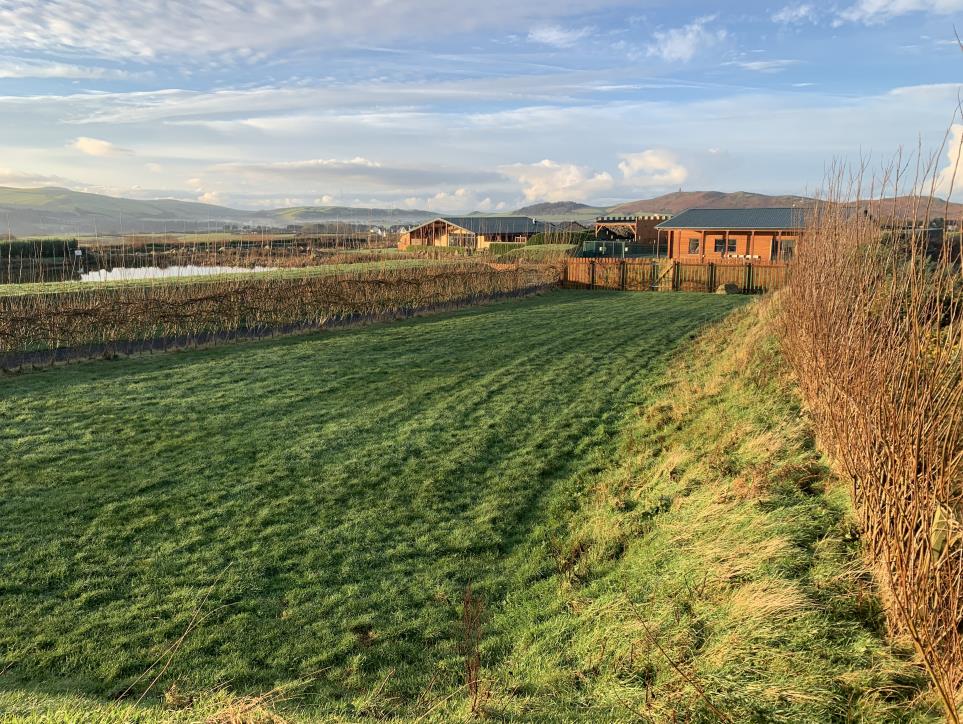 A photograph showing a grassy field in the foreground with a wooden fence, leading to wooden buildings in the distance set against a rural landscape with hills.