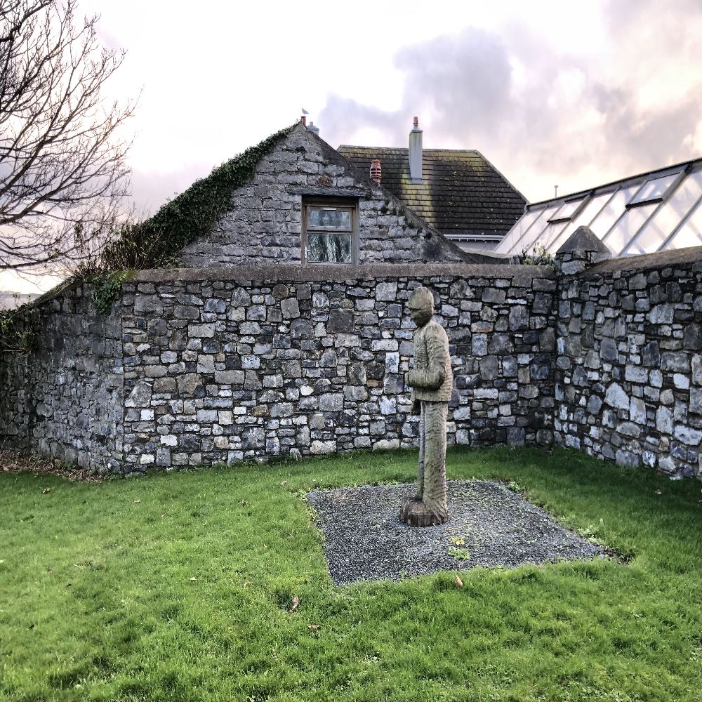 A photograph showing a stone garden wall, a wooden sculpture in a grassy area, and a stone building with a window in the background.