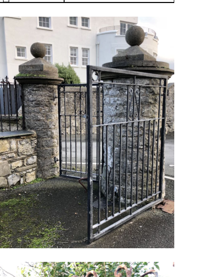 A photograph showing a black metal gate between stone gateposts with spherical finials, in front of a white building.