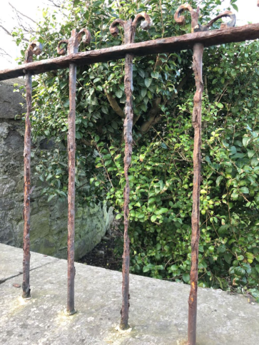 A close-up photograph of rusty metal railings with decorative scrollwork, set against a stone wall and dense green foliage.