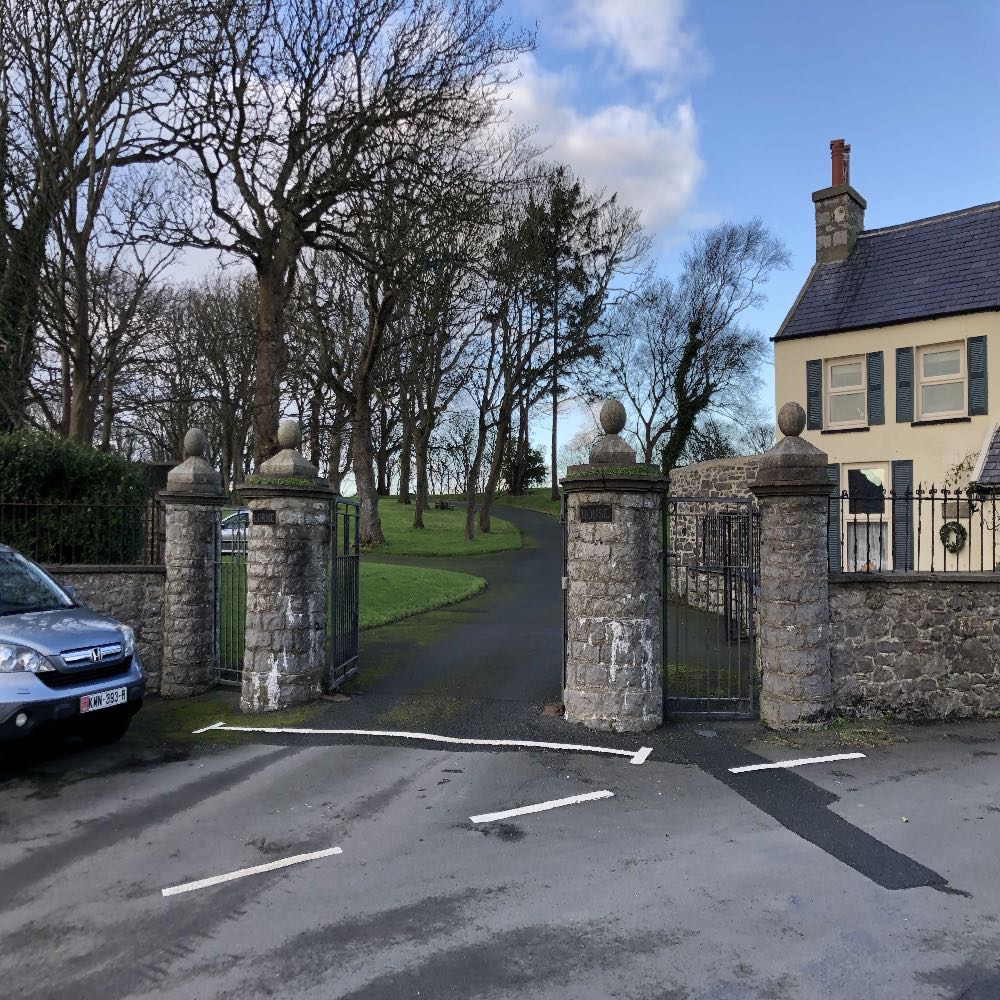 A street-level photograph showing the entrance to a property featuring stone pillars with metal gates and a driveway leading uphill. A cream-colored house is visible on the right side of the frame.