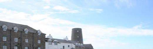 A photograph showing a large building with a slate roof and dormer windows alongside a historic cylindrical tower structure under a blue sky.