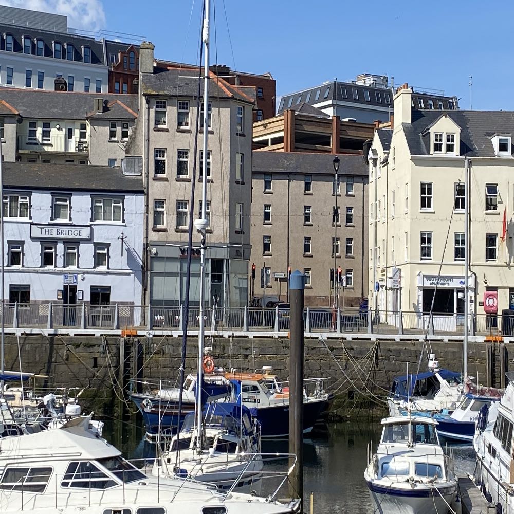 A photograph showing a harbor scene with boats moored in the foreground and a row of multi-story buildings along the waterfront quay in the background.