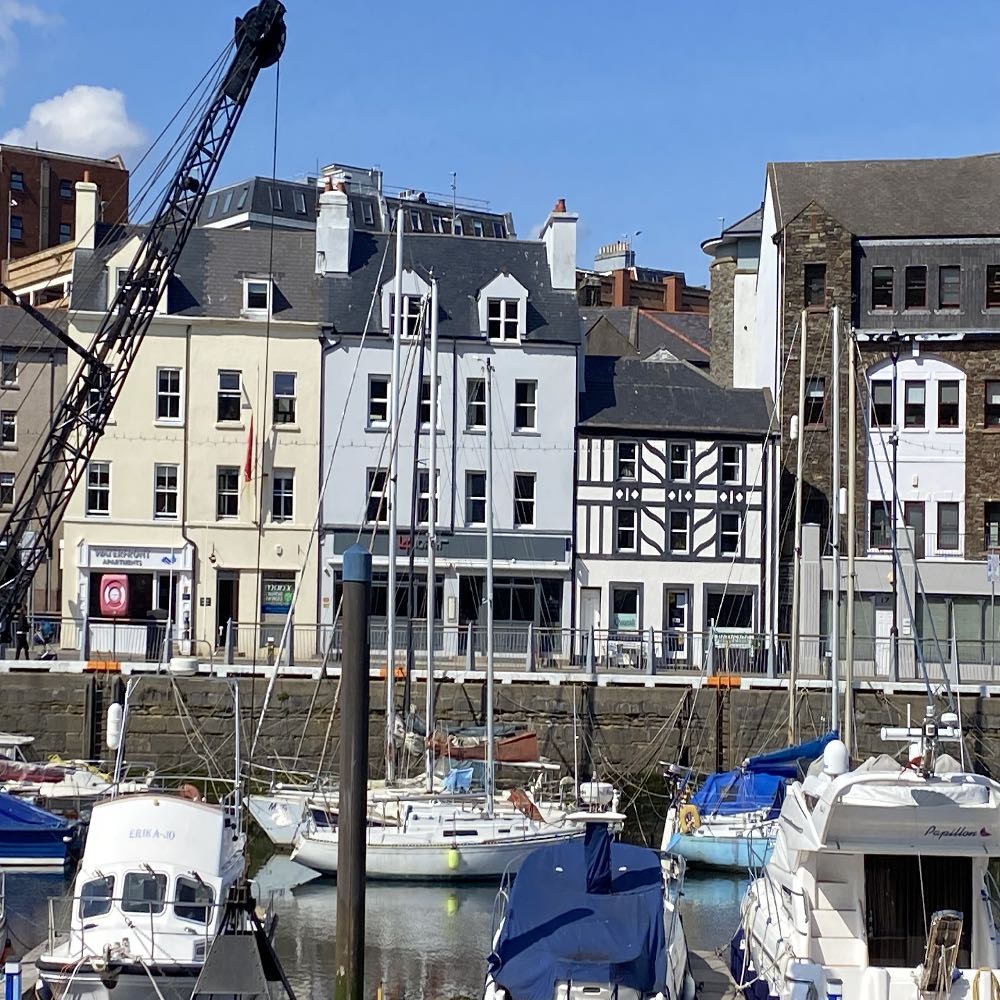 A photograph showing a harbor scene with boats moored in the foreground and a row of mixed-use buildings along the waterfront in the background.