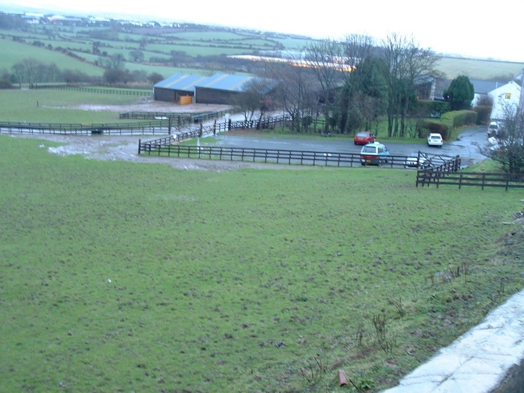 A photograph of a rural site featuring a large agricultural building, a paved area with parked cars, and wooden fencing.