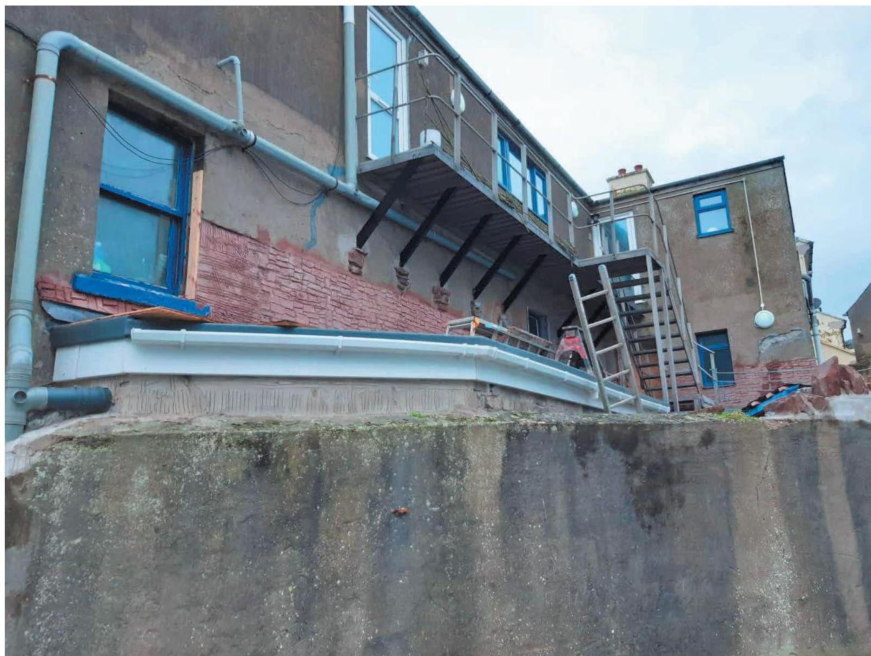 A low-angle photograph of a building's rear elevation undergoing renovation, showing a new first-floor extension with a metal staircase and balcony.