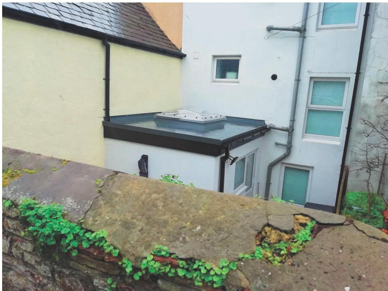 A photograph showing a flat roof extension with a skylight attached to a white building, viewed over a weathered stone boundary wall.