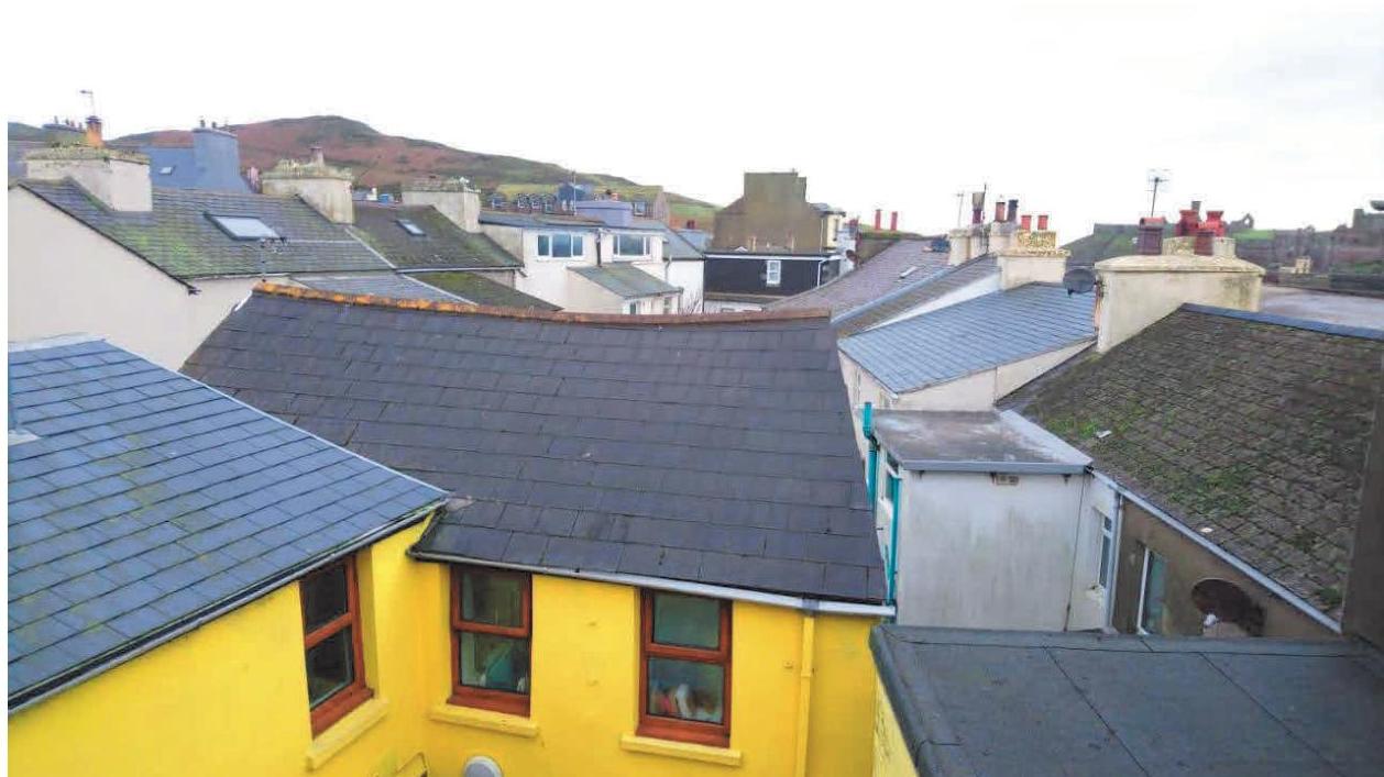 A high-angle photograph showing a cluster of slate rooftops in a town setting, featuring a yellow building in the foreground and hills in the distance.