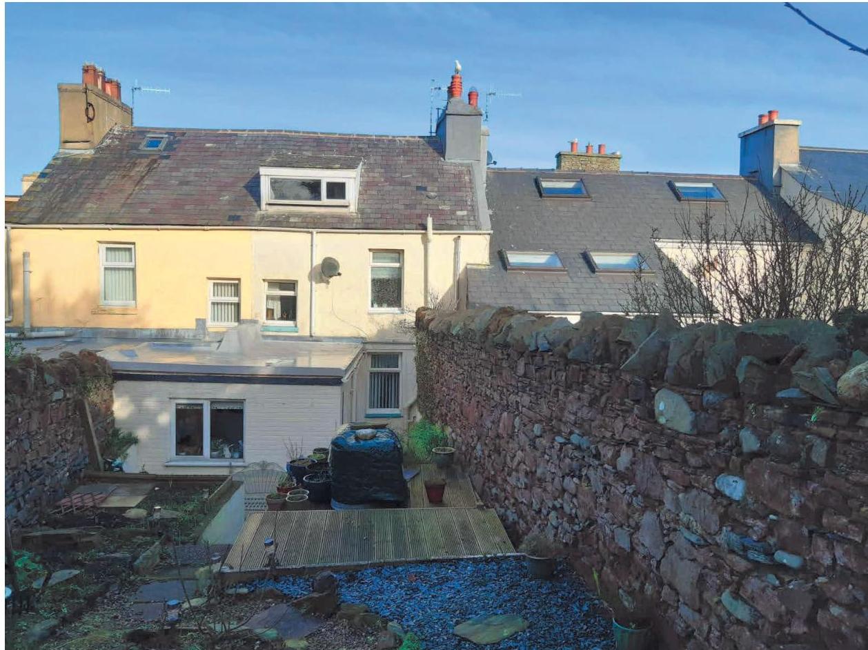 A photograph showing the rear elevation of a terraced house with an existing single-story flat-roofed extension and a garden enclosed by a high stone wall.