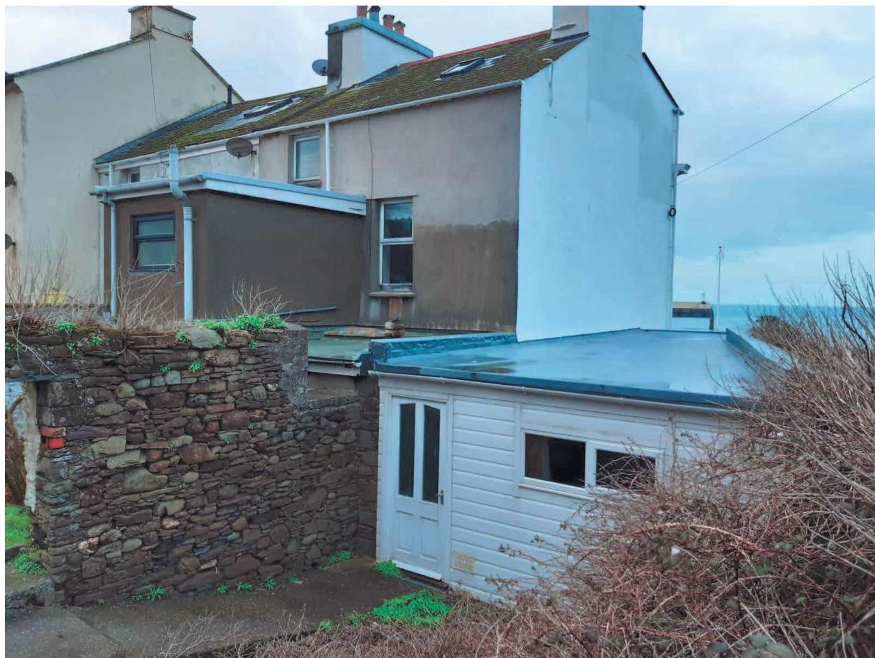 A photograph showing the rear elevation of a residential property with a stone boundary wall in the foreground and the sea visible in the background.