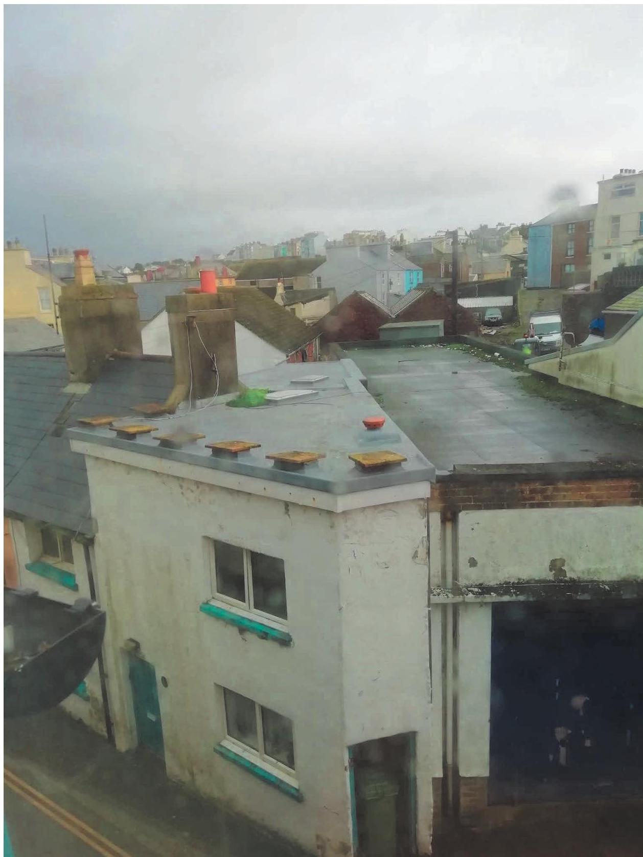 A high-angle photograph showing the flat roof of a white residential building with chimney stacks, overlooking a dense urban area with other rooftops and parked cars in the background.