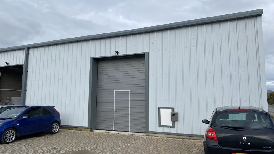 A photograph showing the exterior of a white industrial building with a large grey roller shutter door and a pedestrian door. Two cars are parked on the paved forecourt in front of the structure.