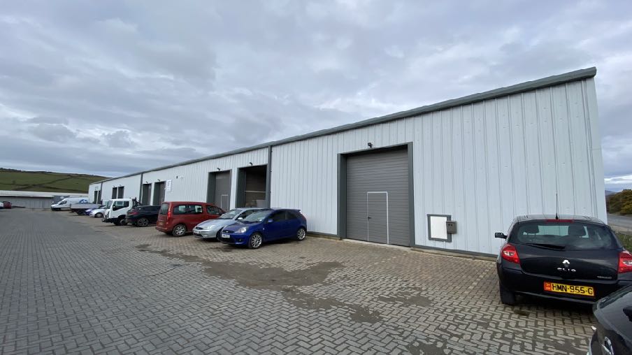 A photograph showing the exterior of a long, white industrial unit with roller shutter doors and several cars parked on a paved forecourt.