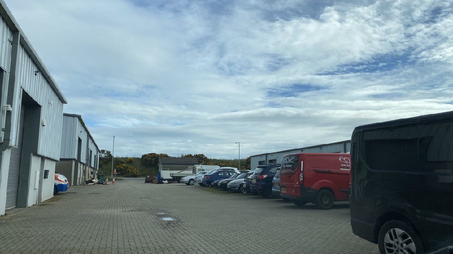 A photograph showing a paved industrial yard with white corrugated metal buildings on the left and parked commercial vehicles.