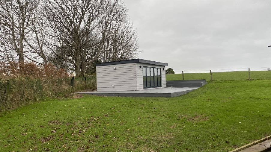 A photograph showing a modern white garden room or office with a flat roof and large glass doors situated on a raised grey deck. The structure is located in a grassy, rural field with trees and a fence in the background.