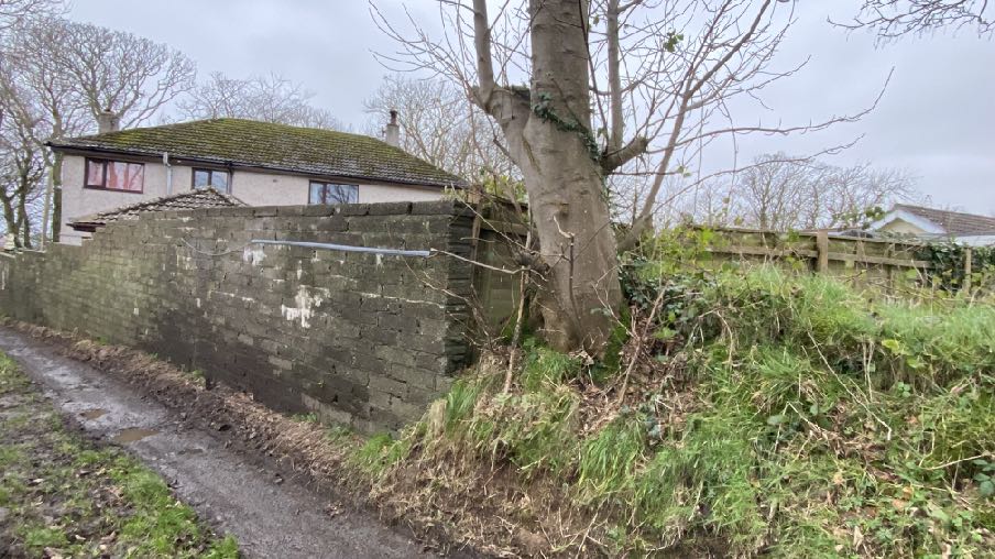 A photograph showing a rural property with a stone boundary wall, a large tree, and a house in the background.