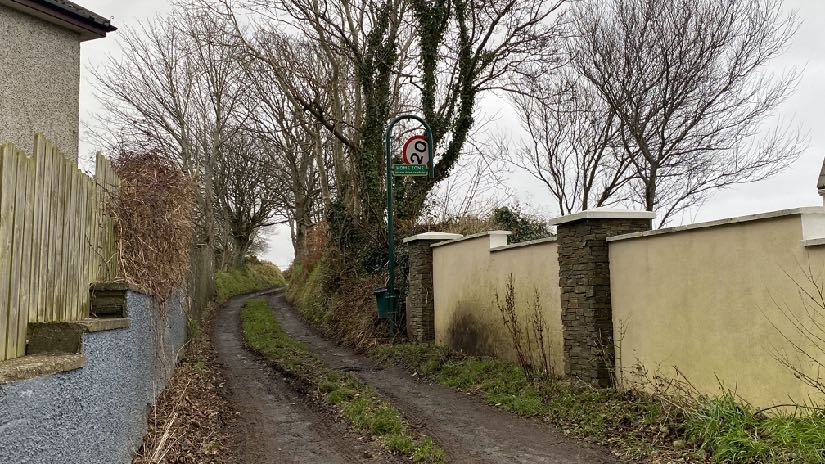 A photograph showing a narrow rural lane with a wooden fence on the left and a cream boundary wall on the right.