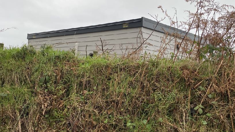 A photograph showing a single-story garden room or office building with light cladding and a dark roof, situated on a grassy slope with overgrown vegetation in the foreground.
