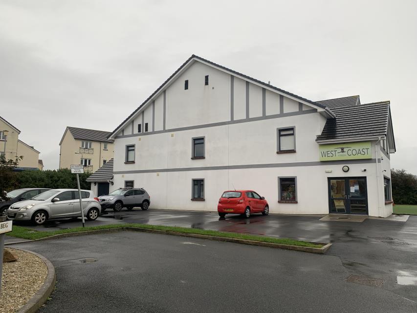 A street-level photograph of a white, two-story commercial building with grey trim and a 'West Coast' sign. Several cars are parked in a wet tarmac area in front of the property.