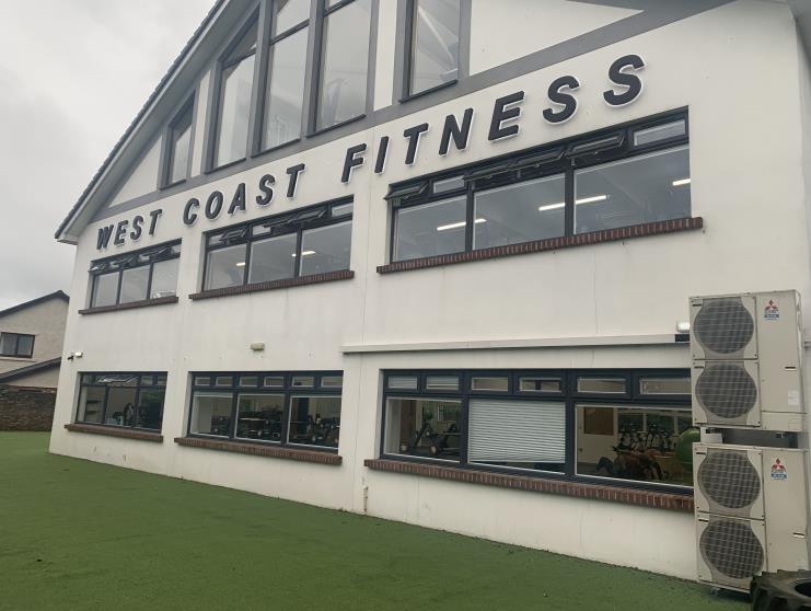 A photograph of the exterior facade of a white two-story commercial building labeled 'WEST COAST FITNESS' with large windows and air conditioning units.
