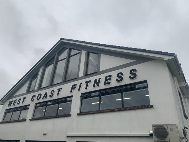 A low-angle exterior photograph of a white commercial building featuring large windows and 'WEST COAST FITNESS' signage.