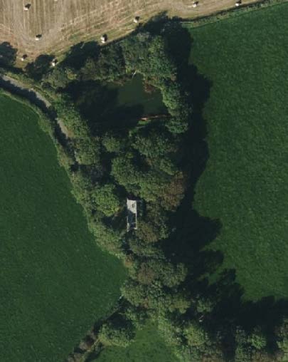 An aerial view of a rural site featuring a building surrounded by dense trees and a small pond, situated between green agricultural fields.