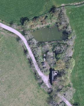 An aerial view of a rural site showing a building complex next to a pond, surrounded by green fields and an access road.