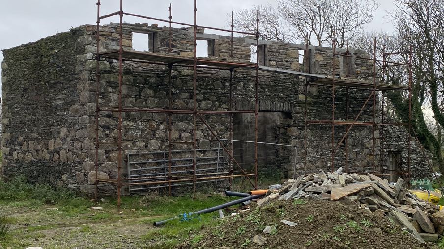 A photograph showing a large stone building, likely a former mill, covered in scaffolding during a renovation project. Construction debris and pipes are visible in the foreground.
