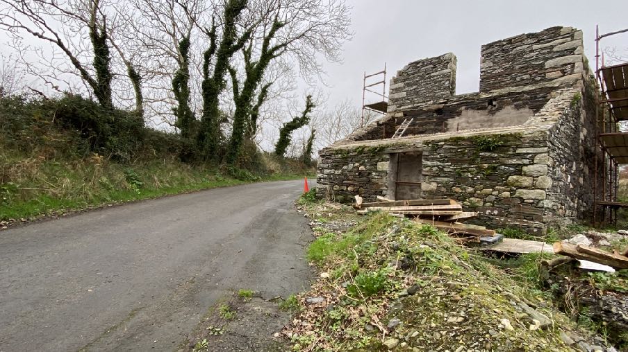 A photograph showing a stone mill building under renovation with scaffolding, situated next to a rural road.
