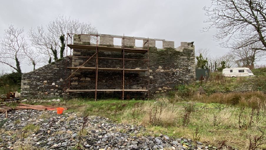 A photograph showing a stone building under renovation with scaffolding erected on the upper wall section. The foreground features a gravel area and the background includes vegetation and a caravan.