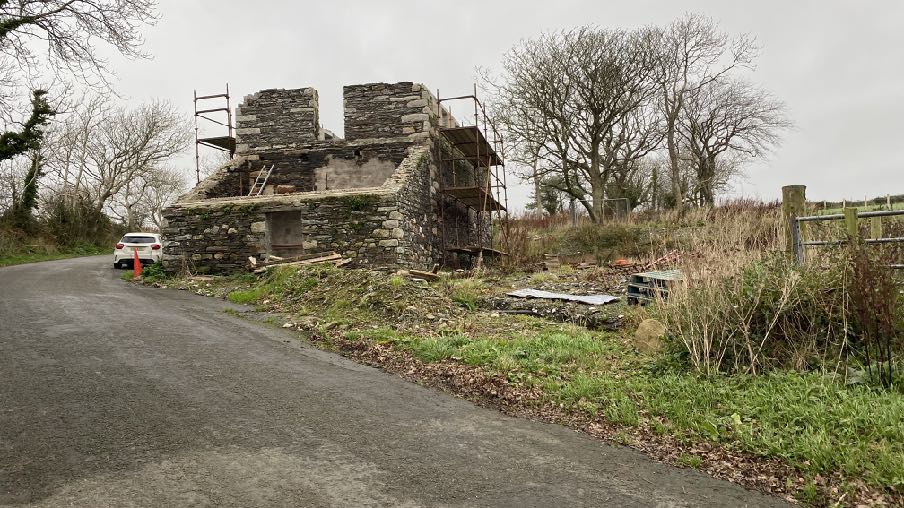 A photograph showing a stone mill building under construction with scaffolding, located beside a paved road in a rural setting.