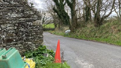 A street-level photograph showing a paved road curving past a rough stone wall on the left, with trees and vegetation in the background and a traffic cone in the foreground.