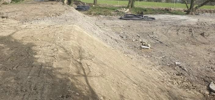 A photograph showing a construction site with earthworks, a mound of sand or soil, and black drainage pipes lying on the ground near trees.