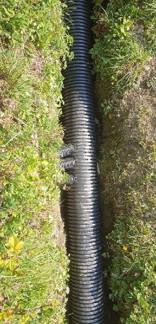 A vertical photograph showing a black corrugated drainage pipe installed in a grassy trench, with smaller branch pipes visible.