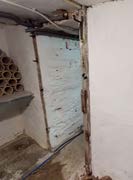 Interior photograph of a room undergoing renovation, showing exposed pipes, unfinished walls, and a doorway covered with a blue board.