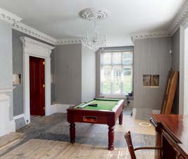 A photograph of a residential interior room featuring a pool table, a large window, and traditional architectural details like cornices and a fireplace surround.