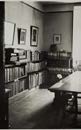 A black and white photograph showing an interior room with built-in bookshelves and framed artwork on the walls, likely a study or library.
