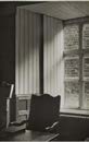 A black and white interior photograph showing a window with vertical blinds and a chair in the foreground.
