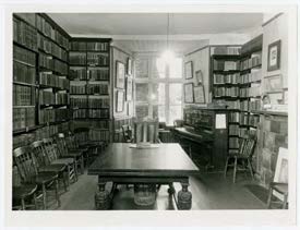 A black and white photograph showing the interior of a historic room, likely a library or study, featuring floor-to-ceiling bookshelves, a large table, and a piano.