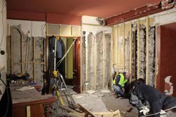 The image shows the interior of a building undergoing renovation with walls stripped to exposed stone and timber, and workers present on site.