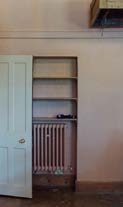 Interior photograph showing a white panelled door, a built-in shelving unit, and a radiator against a cream wall.