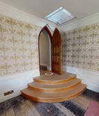Interior photograph showing a curved wooden staircase leading to an arched wooden screen, featuring patterned wallpaper and a skylight.