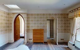 Interior photograph of a room with patterned wallpaper, wooden floors, and an arched doorway, likely showing existing conditions for a restoration project.