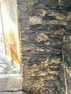 A close-up photograph of a rough, irregular stone wall, likely an interior detail of a historic building, showing masonry texture and a wooden beam.