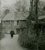 A black and white photograph showing a traditional stone cottage with a person walking on a path in the foreground.