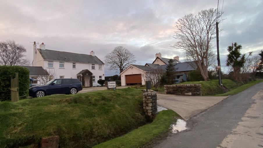 A street-level photograph showing a white detached house named Poorna Cottage and a separate garage building with a brown door, situated on a grassy slope next to a road.