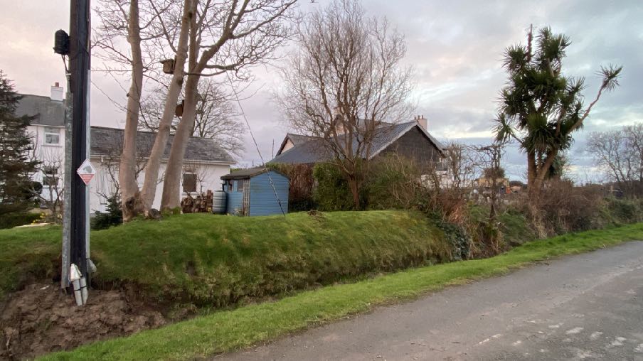 A street-level photograph showing a blue wooden outbuilding or shed situated on a grassy bank next to a road, with residential houses and trees in the background.