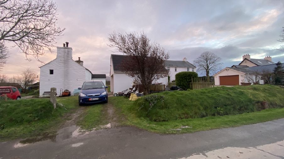 A street-level photograph showing a rural residential scene with white-washed buildings, a driveway with a parked car, and a grassy verge.