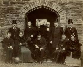 A vintage black and white photograph showing a group of formally dressed people posing in front of a stone building with a large arched doorway.