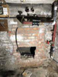 A grainy photograph showing an interior brick wall with exposed pipes and a dark rectangular opening, likely a fireplace or stove recess.